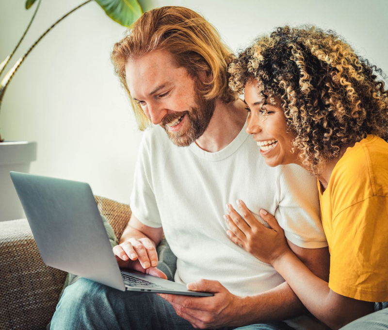 Multiracial young couple watching computer laptop sitting on the sofa at home - Happy diverse husband and wife using pc online services - Technology life style concept